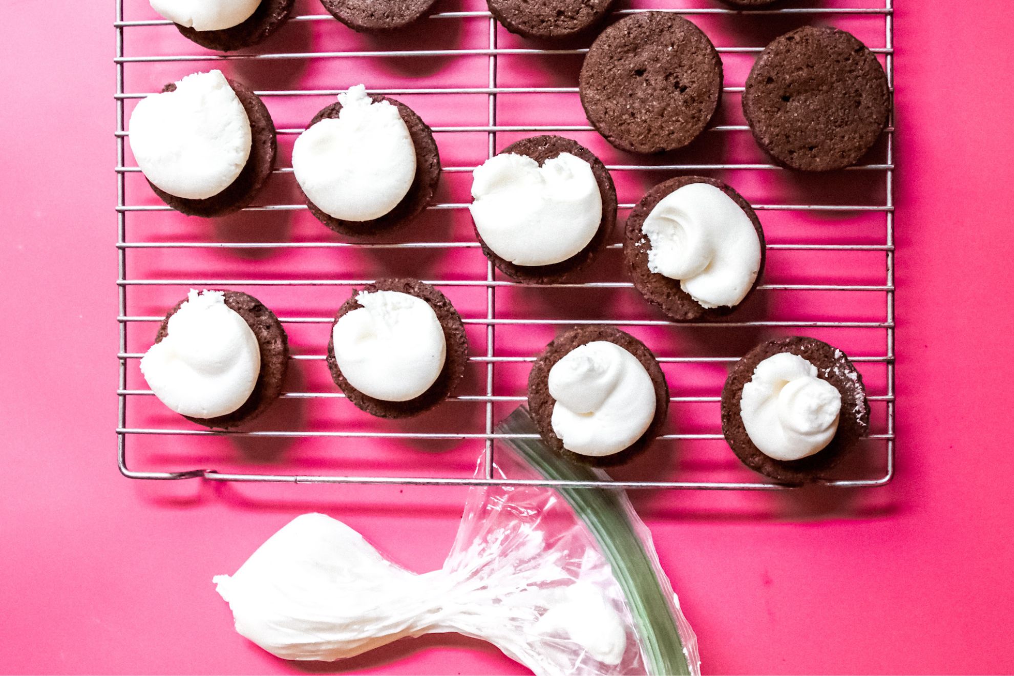 This is an overhead horizontal image of a silver cooling rack on a dark pink surface. On the cooling rack are chocolate circle cookies. Half of the cookies have vanilla icing piped out on the center. To the bottom center of the image of a plastic baggie with vanilla frosting in it.