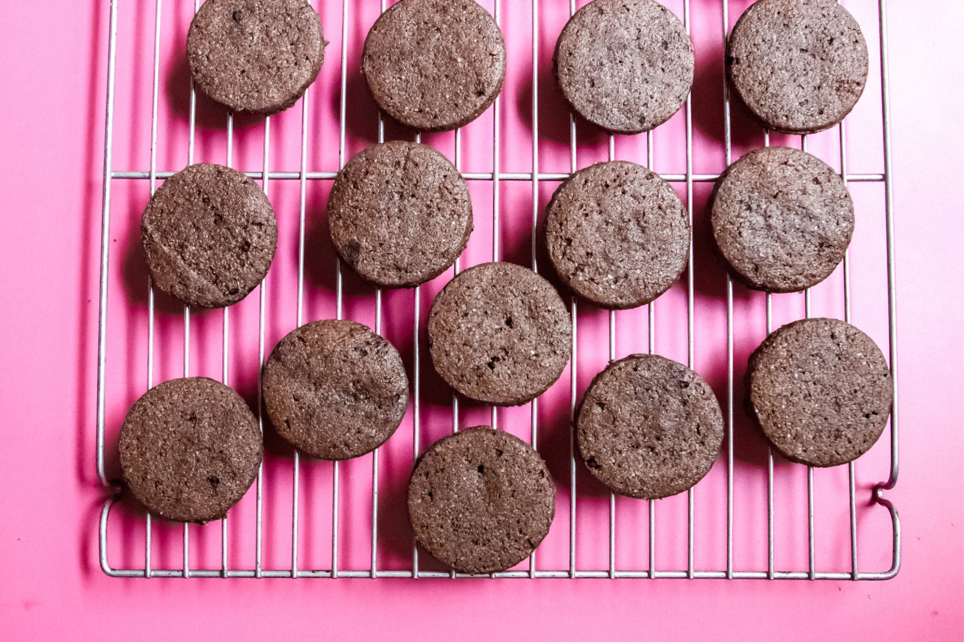 This is an overhead horizontal image of a silver cooling rack on a dark pink surface. On the cooling rack are chocolate circle cookies.