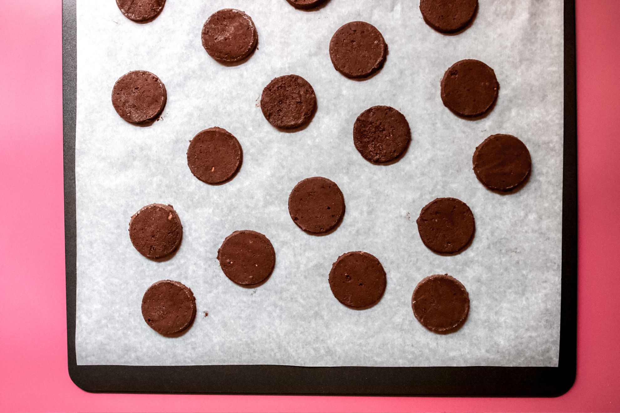 This is an overhead horizontal image of a baking sheet lined with white parchment paper. On the parchment paper are raw flat, chocolate circle cookies. The baking sheet sits on a dark pink surface.