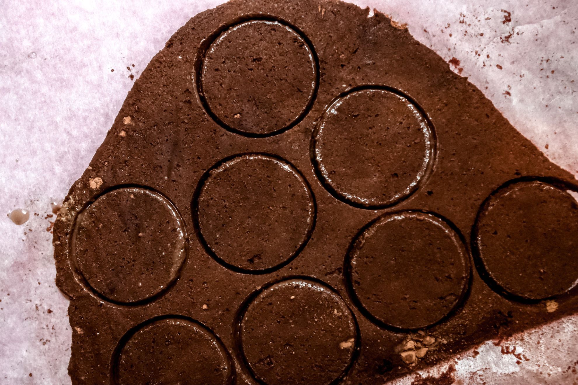 This is a closeup of chocolate cookie batter rolled out on a white piece of parchment paper. Circles are pressed and cut from the rolled cookie dough but have not yet been separated.