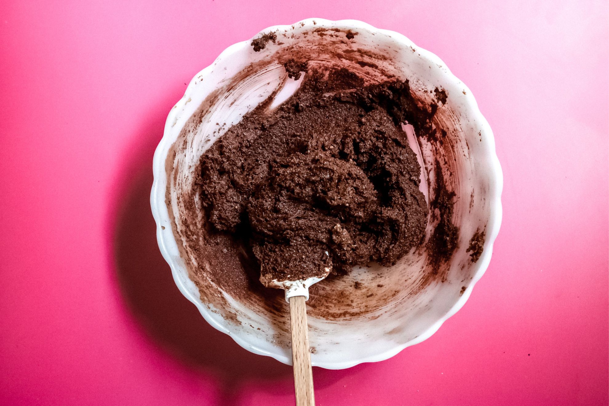 This is an overhead horizontal image of a white milk glass scalloped bowl on a dark pink surface. In the bowl is chocolate batter and a white rubber spatula with a wooden handle leaning against the side and pointing to the bottom center of the image.