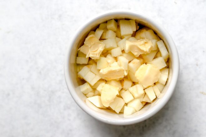 This is a horizontal overhead image of a small bowl of peeled and cubed apples with small dollops of butter on top. The white ramekin sits on a light grey/white surface.