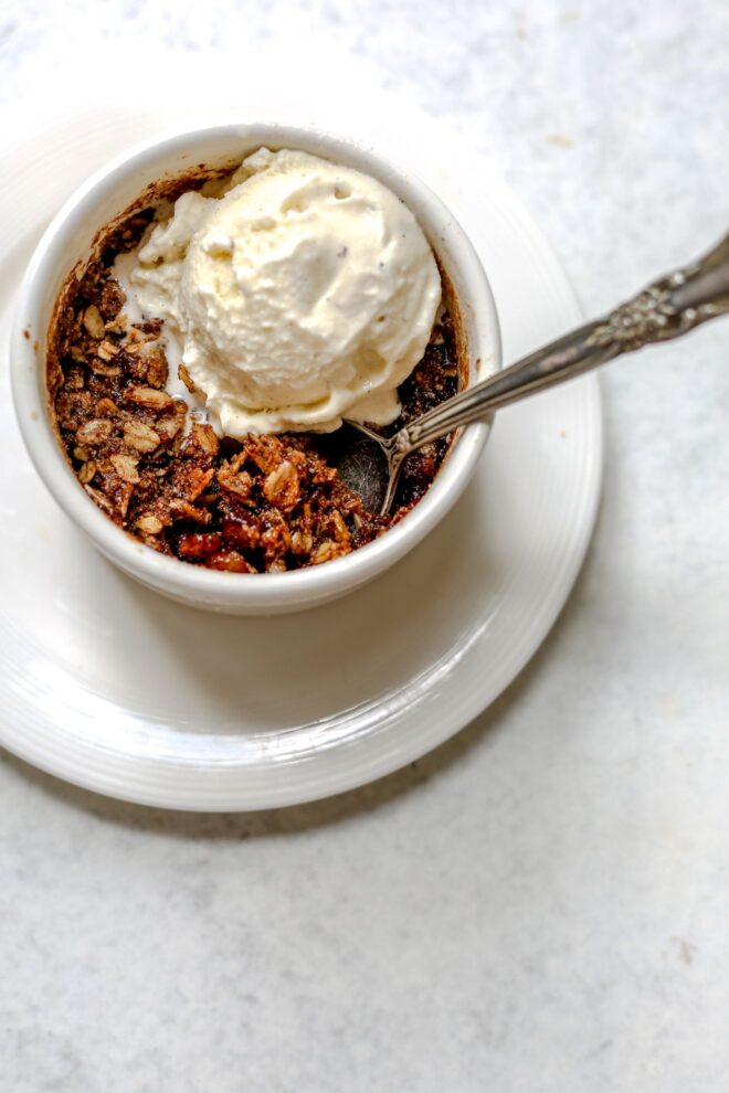 This is an overhead vertical image of a small white ramekin with a baked apple oat crisp in it. A big scoop of vanilla ice cream is on top of the crisp. The ramekin is on a small white plate. A silver spoon has dipped into the apple and oat crisp and leaning against the side of the ramekin with the silver handle pointing to the right side of the image. The plate sits on a light grey surface.