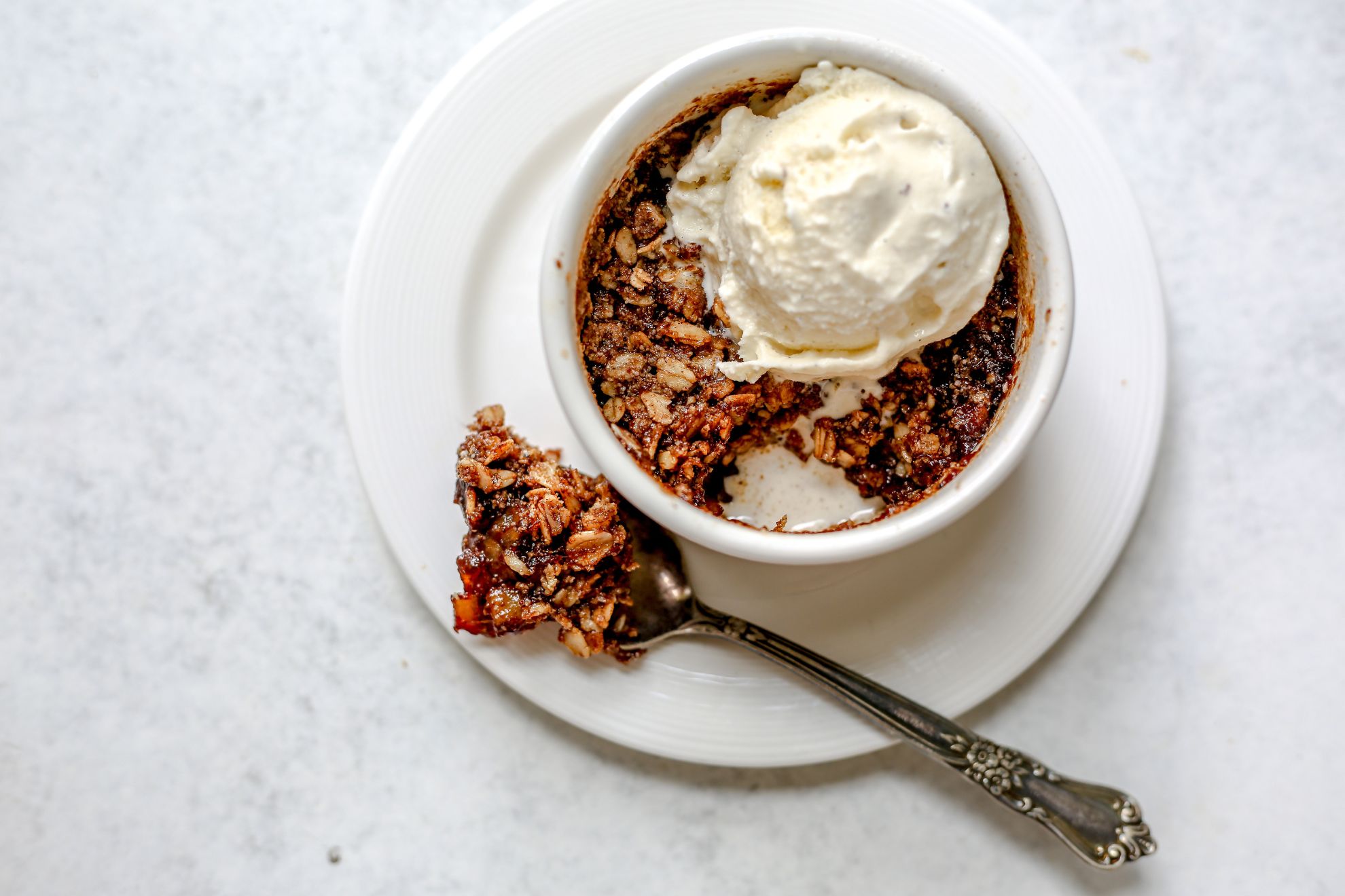 This is an overhead horizontal image of a small white ramekin with a baked apple oat crisp in it. A big scoop of vanilla ice cream is on top of the crisp. The ramekin is on a small white plate. A silver spoon has scooped a bite of the apple crisp out and is lying next to the ramekin on the plate. The plate sits on a light grey surface.