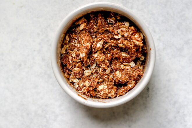This is an overhead horizontal image of a small white ramekin with an oat, coconut sugar crumble on top. The ramekin sits on a light grey white surface.