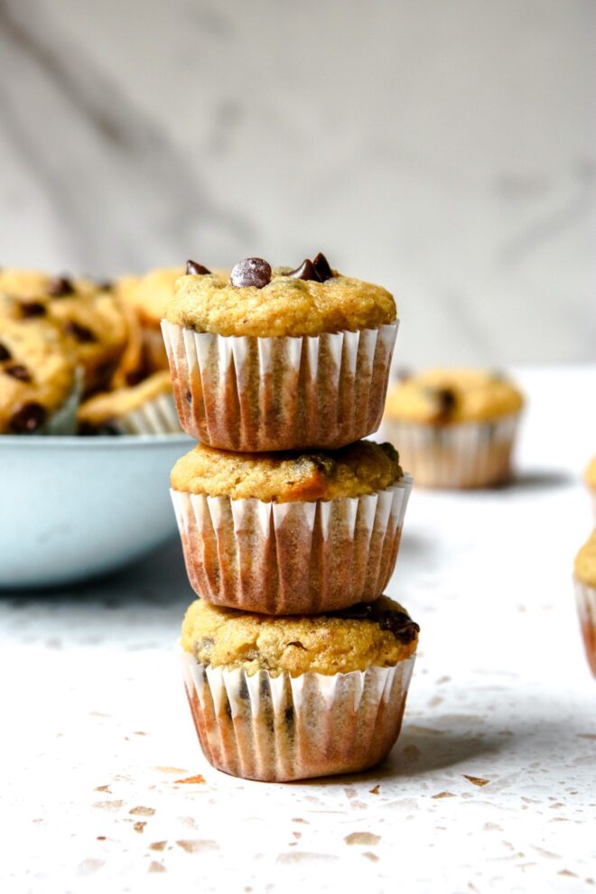 This is a vertical image looking at a stack of three muffins from the side. The muffins sit on a white and beige terrazzo surface. Behind the stack of muffins is a light blue bowl filled with muffins and more muffins are on the counter, blurred in the background behind the stack.