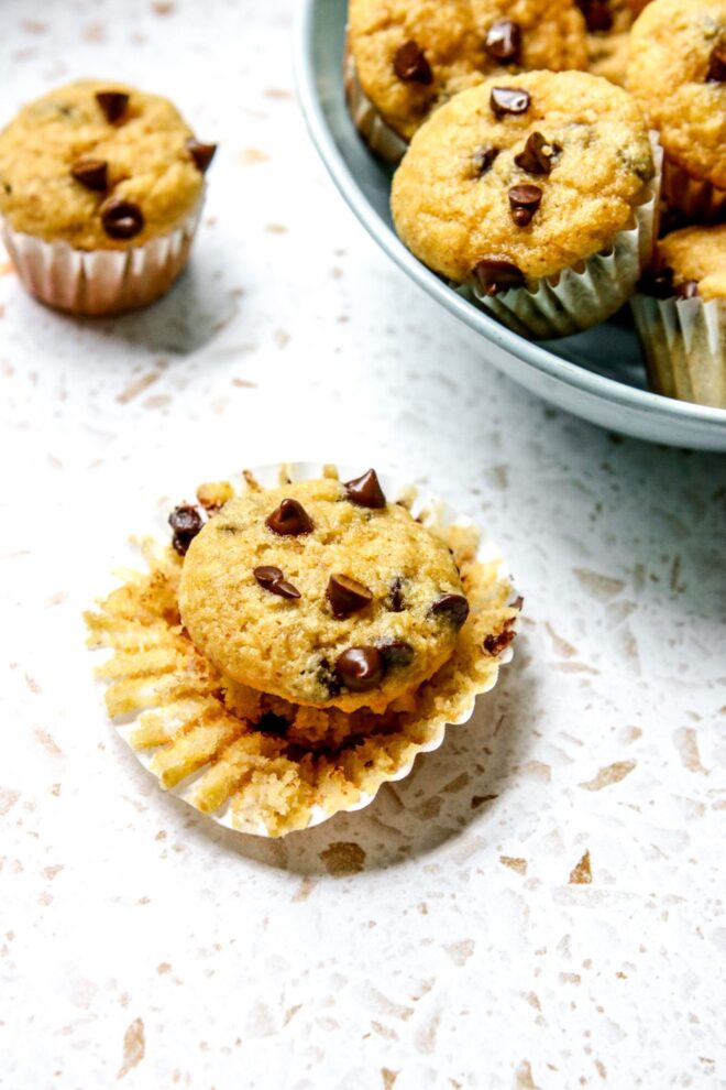 This is a vertical image looking at a mini muffin from the side. The parchment liner is pulled away from a chocolate chip mini muffin. It sits on a white and beige terrazzo surface. To the top right of the image is a light blue bowl with more mini muffins and another mini muffin is on the surface to the top left corner of the image.