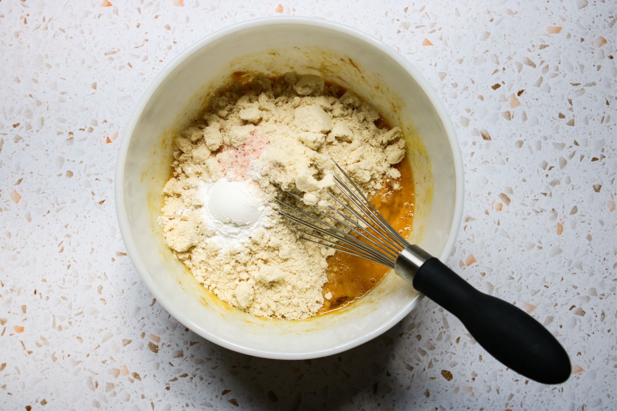 This is an overhead horizontal image of a large white bowl with peanut butter colored mixture in it. On top of the wet mixture is blanched almond flour, a white power and pink salt. The bowl is centered in the middle of the image on a white, beige and peach terrazzo surface. A whisk with a black rubber handle is in the bowl with the handle leaning against the side of the bowl and pointing to the bottom right corner of the image.