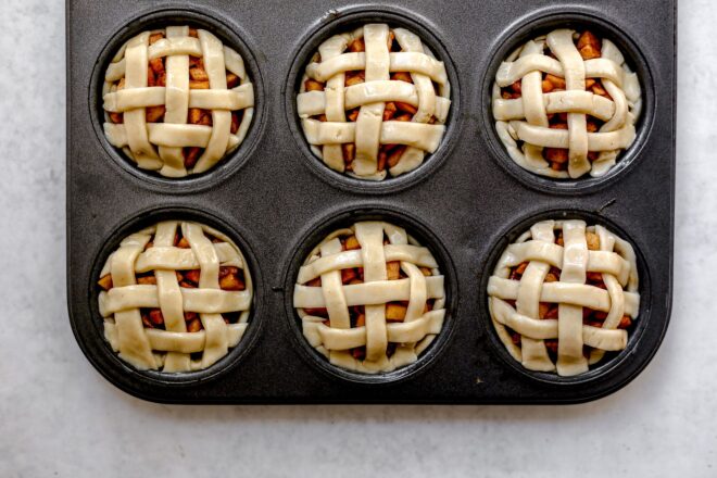 This is an overhead horizontal image of a six cup muffin tin with raw lattice pie crust topping over diced apples. The muffin tin sits on a light grey surface.