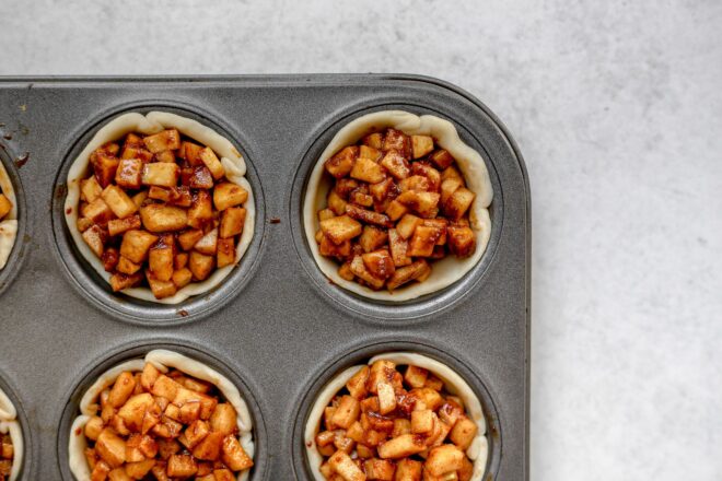 This is an overhead horizontal image of a muffin tin with pie crust pressed into the cups and up the sides. Wet cinnamon coated apple pieces are in the dough cups. The muffin tin sits on a light grey surface.