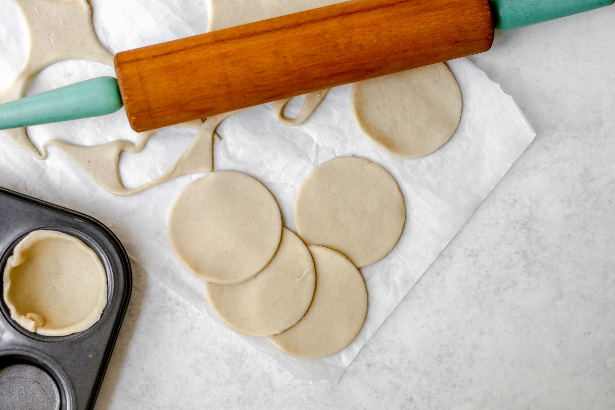 This is a horizontal overhead image of pie dough cut in circles. The dough circles are on a white piece of parchment paper with the leftover dough to the top left, a wood rolling pin with teal handles to the top center of the image, and the corner of a muffin tin with a pie dough round pressed into the cup and up the sides.