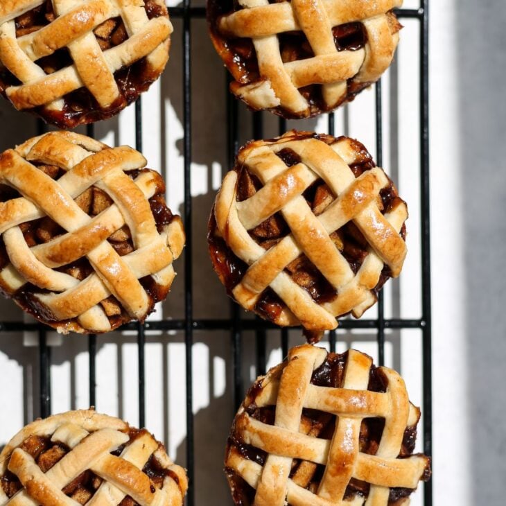 This is a vertical overhead image of a cooling rack with six mini lattice topped pies on it. The cooling rack is on a white surface.