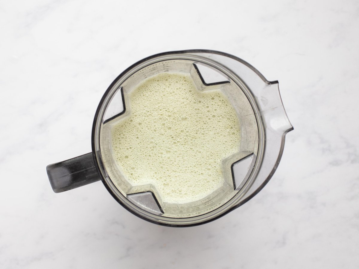 This is an overhead horizontal image looking into a blender with a bubbly pale green liquid in it. The blender sits on a white marble surface.