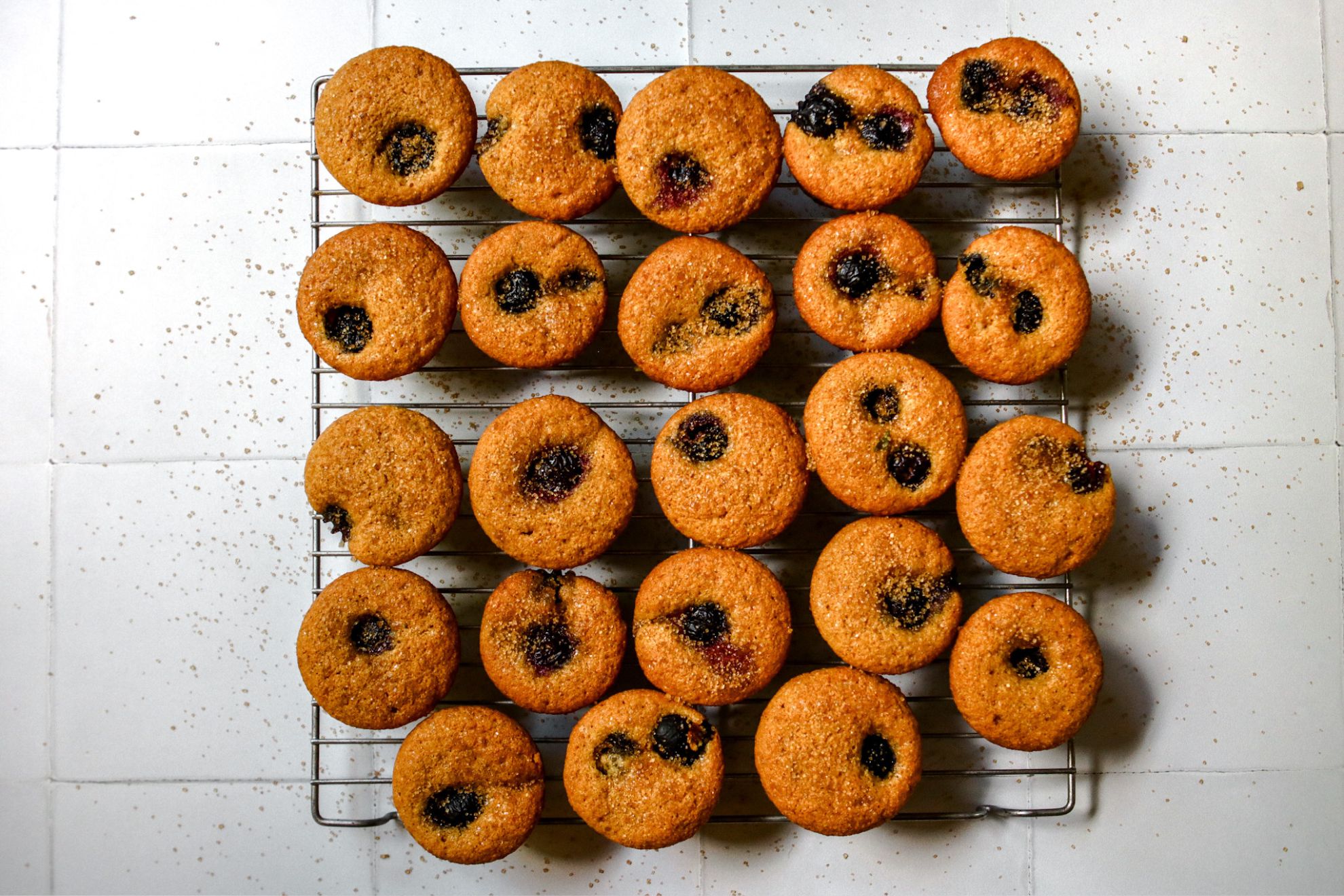 This is an overhead horizontal image of small muffins on a silver cooling rack. The image shows the entire wire rack on a white tiled surface. The muffins are studded with one or two blueberries and sprinkled with turbinado sugar. Some sugar has fallen down onto the white surface.