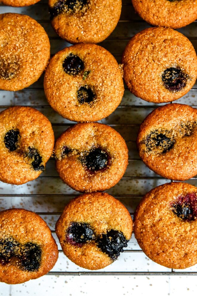 This is an overhead vertical image of small muffins on a silver cooling rack. The muffins are studded with one or two blueberries and sprinkled with turbinado sugar. Some sugar has fallen down onto the white surface. The image is closeup on three lines of muffins on the rack.
