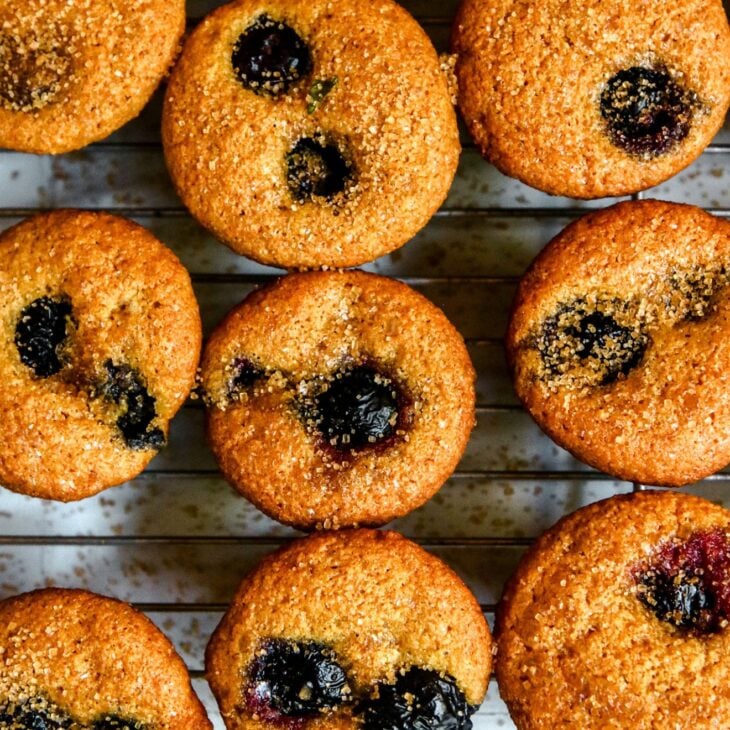 This is an overhead vertical image of small muffins on a silver cooling rack. The muffins are studded with one or two blueberries and sprinkled with turbinado sugar. Some sugar has fallen down onto the white surface. The image is closeup on three lines of muffins on the rack.