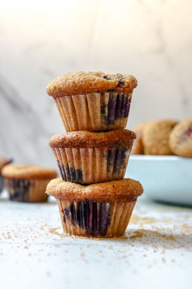 This is a vertical image looking at a stack of three mini blueberry muffins from the side. The stack sits on a white surface with grains of brown sugar scattered around it. The muffins are studded with blueberries under the white translucent muffin liners. Blurred behind the stack is a light blue bowl with more muffins to the right and a couple muffins on the white surface to the left.