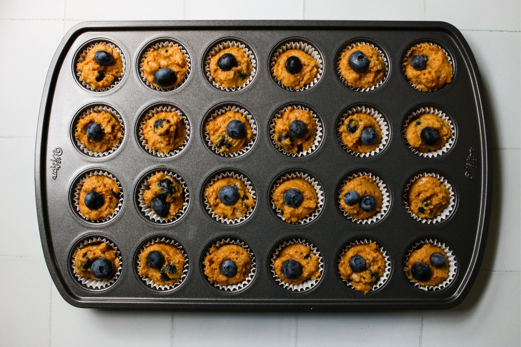 This is an overhead horizontal image of a 24 cup mini muffin tin on a white tile surface. The tin has white muffin liners and batter topped with blueberries.