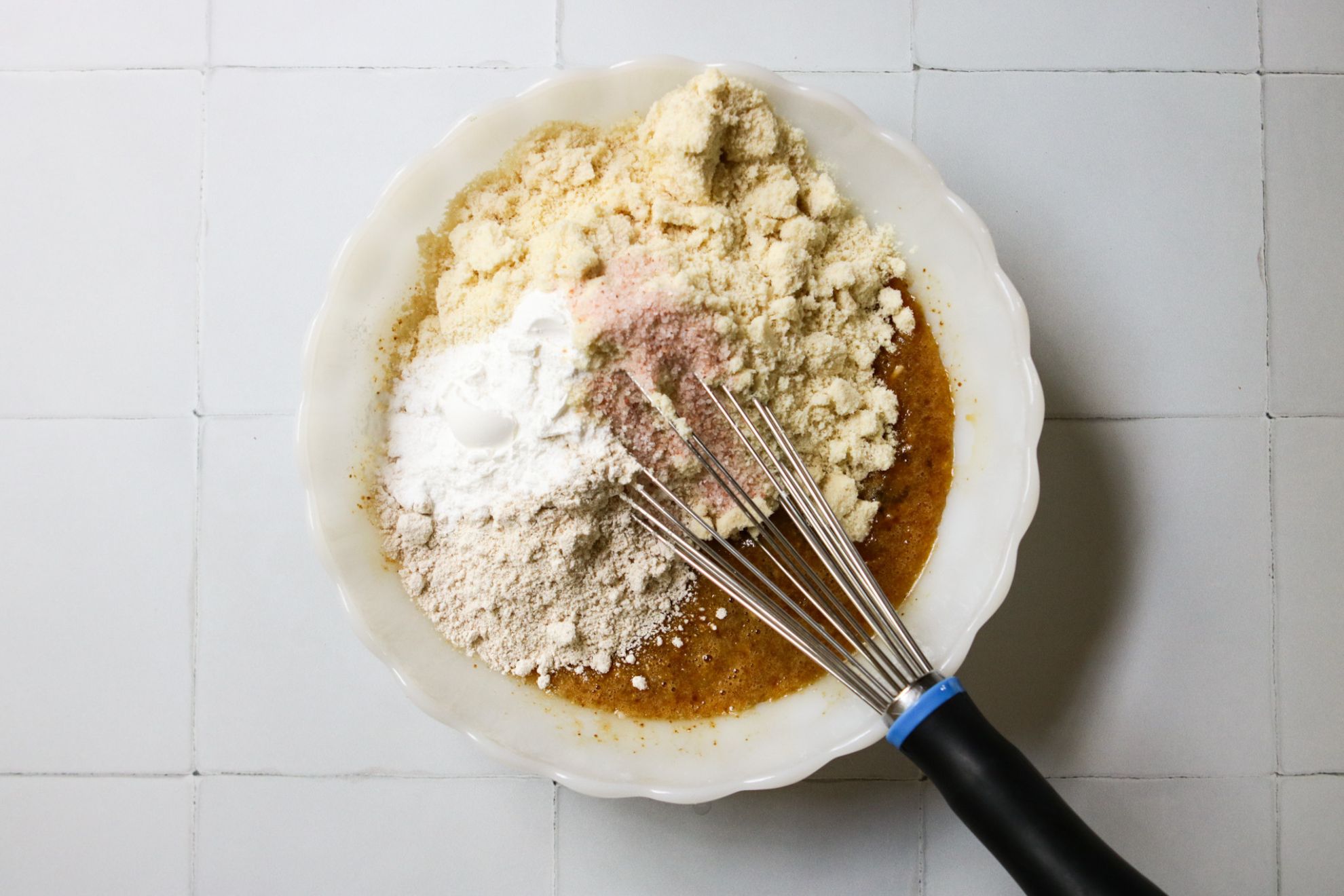 This is an overhead horizontal image of a scalloped milk glass bowl with almond flour, oat flour, salt, and baking powder in it. Underneath the dry ingredients are wet ingredients mixed together. A whisk is in the bowl, leaning against the side with the handle pointing to the bottom right corner of the image. The bowl sits on a white tiled surface.