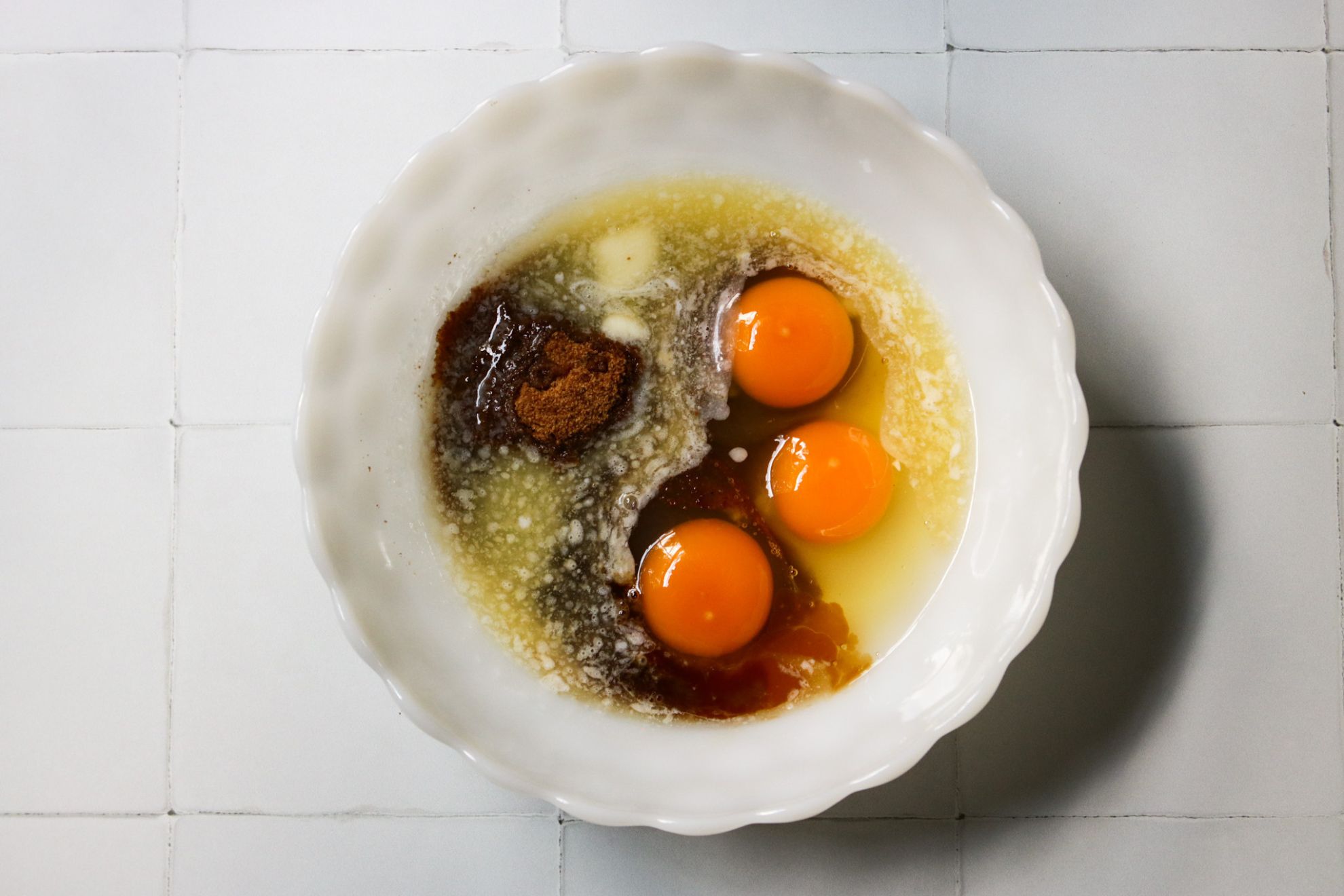 This is an overhead horizontal image of a scalloped milk glass bowl with melted butter, three eggs, and coconut sugar in it. The bowl sits on a white tiled surface.