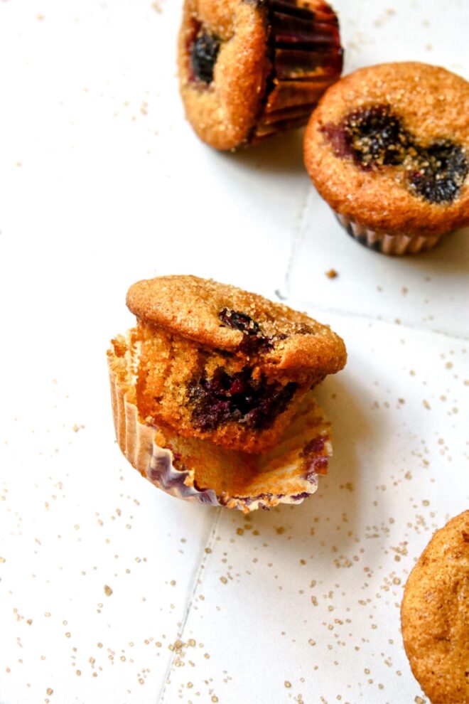 This is a vertical image focusing on a single muffin on a white tiled surface. The muffin has its liner pulled away and is leaning on its side. the muffin has blueberries and is sprinkled with turbinado sugar. More sugar has sprinkled onto the surface. More muffins are peaking in from the bottom right corner and two blurred in the top right corner.