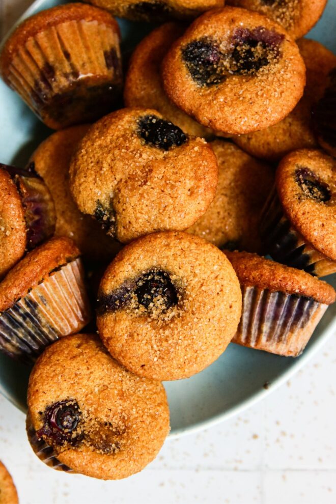 This is an overhead vertical image of a shallow light blue bowl on a white tiled surface. In the bowl are small muffins studded with blueberries and sprinkled with brown sugar. The image is a closeup view of the bowl focusing on the tops of the muffins.