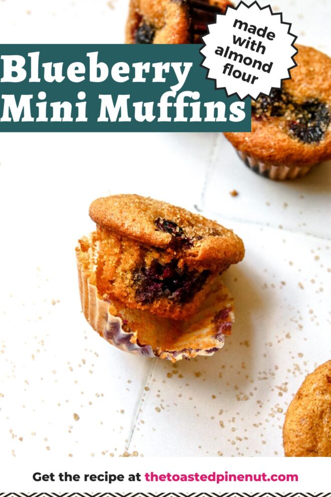 This is a vertical image focusing on a single muffin on a white tiled surface. The muffin has its liner pulled away and is leaning on its side. the muffin has blueberries and is sprinkled with turbinado sugar. More sugar has sprinkled onto the surface. More muffins are peaking in from the bottom right corner and two blurred in the top right corner. Text overlay reads "blueberry mini muffins made with almond flour".