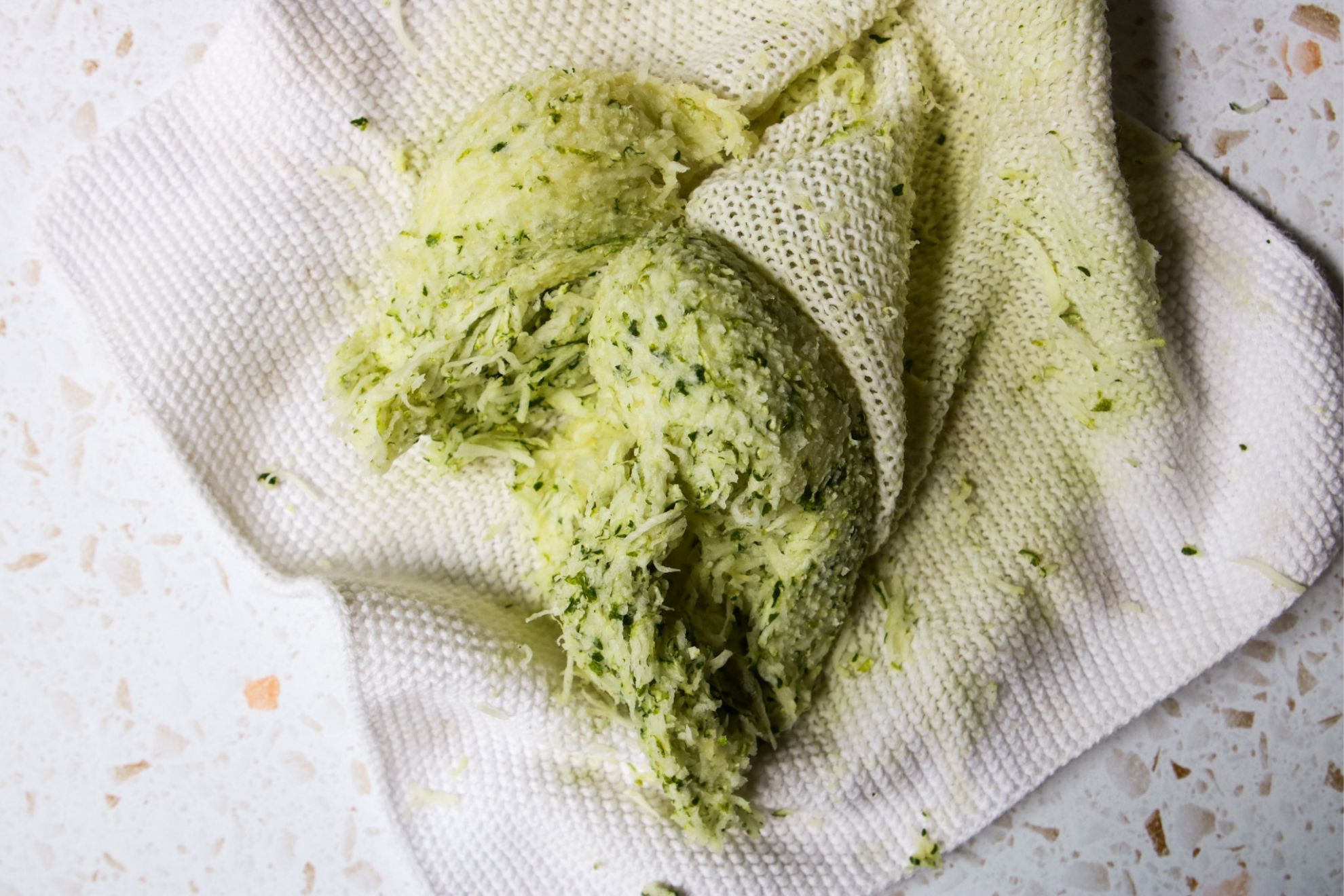 This is an overhead horizontal image of mounds of shredded zucchini on a white knitted towel. The shredded zucchini has been squeezed and so the mounds of zucchini are compressed and the towel has light green stains on it. The towel sits on a white terrazzo surface.