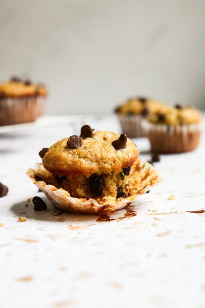 This is a vertical image looking at a muffin from the side, The muffin has chocolate chips on top and the white muffin liner is pulled away from the base. The muffin sits on a white terrazzo surface and more muffins are blurred in the background.