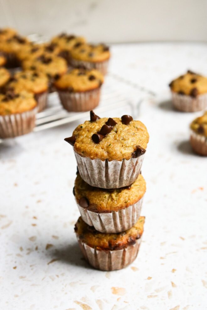 This image looks at a stack of three mini muffins from an angled view. The muffins have white paper liners on them and chocolate chips on top. The stack sits on a white terrazzo surface with more mini muffins blurred in the background.