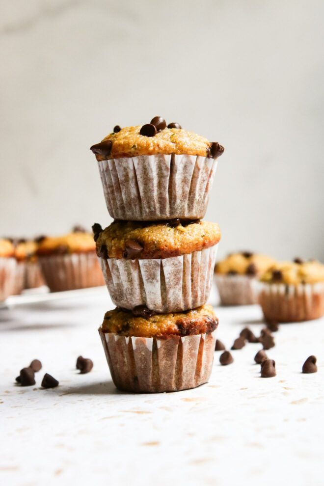 This is a vertical image looking at a stack of three muffins from the side. The muffins have white paper liners on them and chocolate chips on top. The stack sits on a white surface with more mini muffins blurred in the background and mini chocolate chips on the surface scattered around the muffins.
