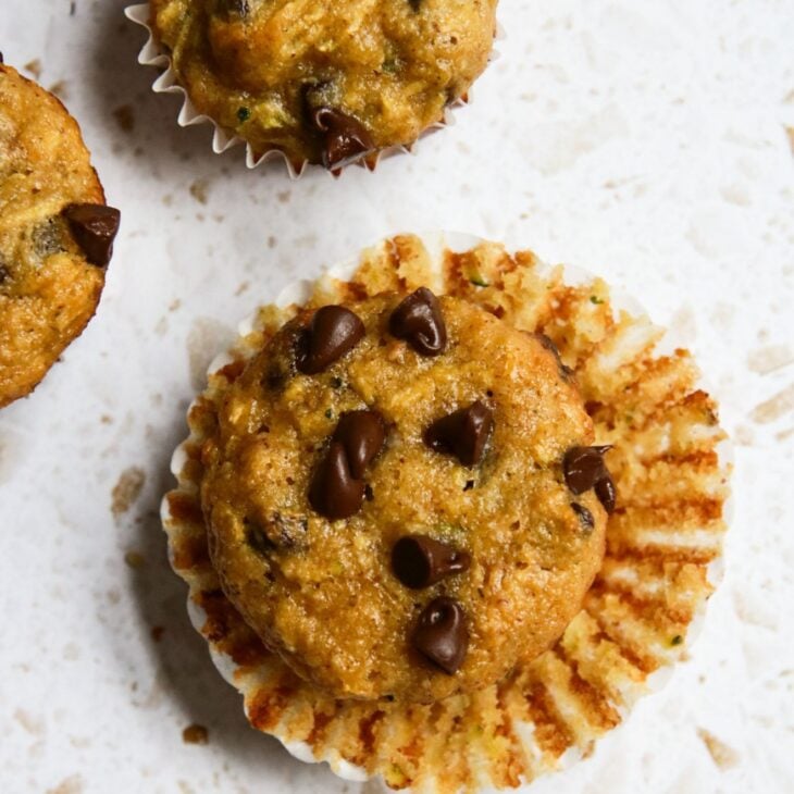 This is a vertical overhead image of a mini muffin with mini chocolate chips on top. The white paper liner is pulled away from the side of the muffin. The muffin sits on a white terrazzo surface. To the top center and left corner of the image are mini muffins
