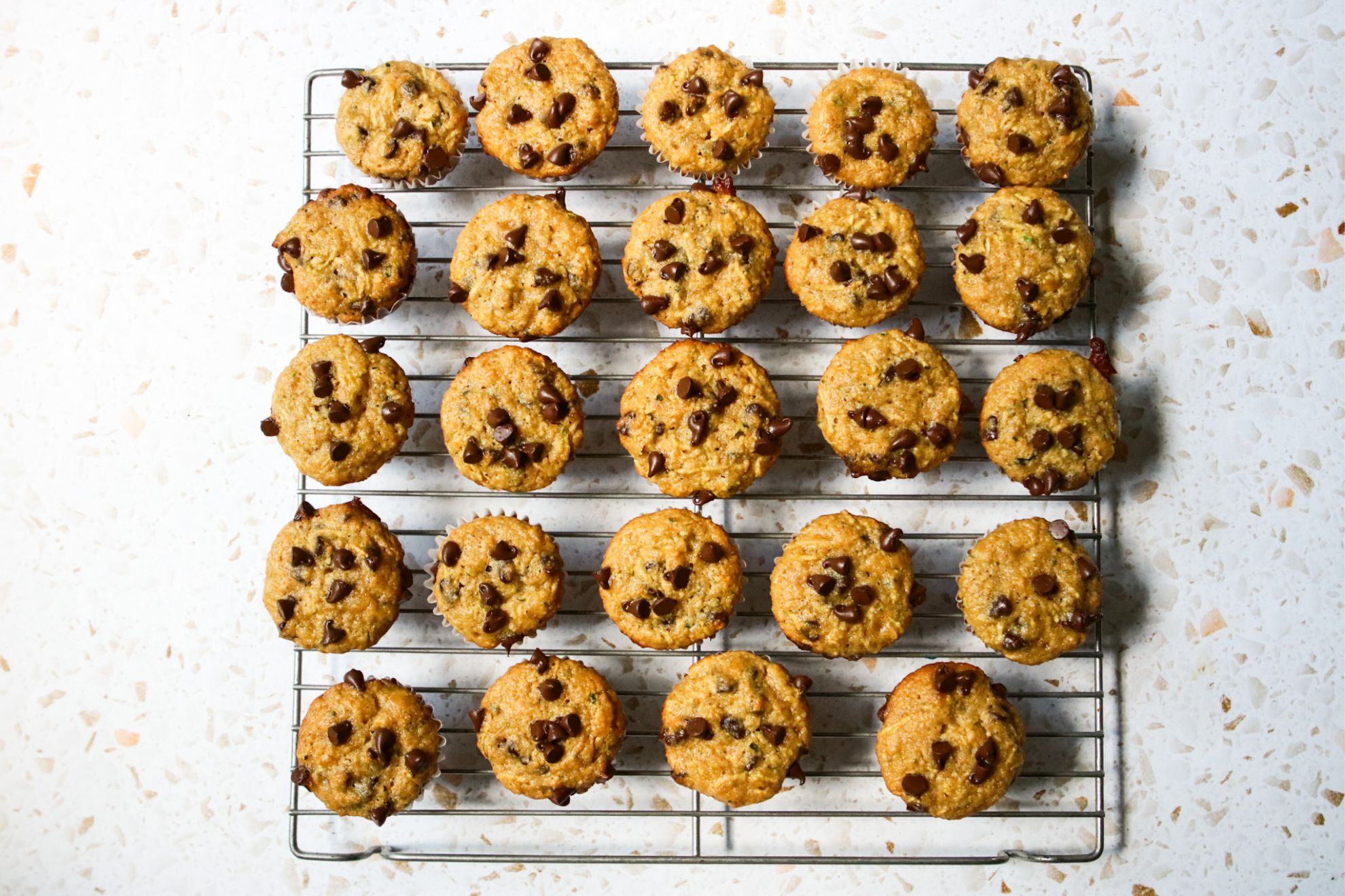 This is an overhead horizontal image of mini muffins on a silver cooling rack. The muffins are topped with mini chocolate chips. The cooling rack sits on a whiter terrazzo surface.