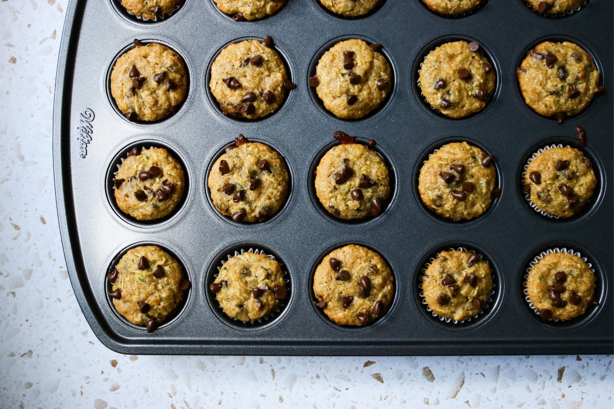 This is an overhead horizontal image of a dark grey mini muffin tin with baked muffins in it. On top of the muffins are mini chocolate chips. The muffin pan sits on a white terrazzo surface.