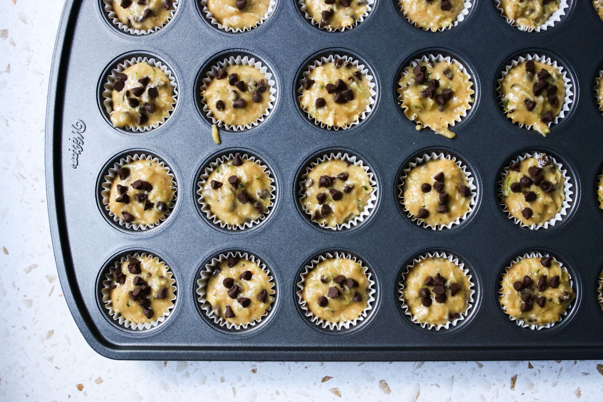 This is an overhead horizontal image of a dark grey mini muffin tin with raw muffin batter in it. On top of the muffins are mini chocolate chips. The muffin pan sits on a white terrazzo surface.