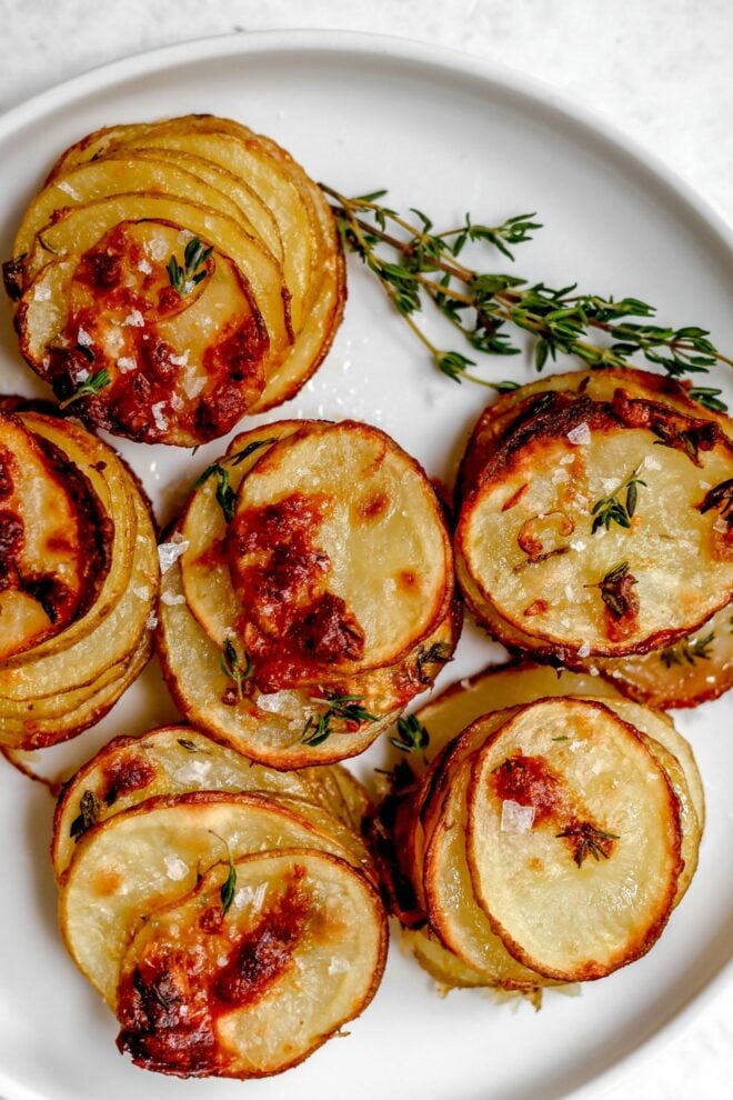 This is an overhead vertical image of sliced stacked white potatoes. The potatoes have golden brown bubbles on them and are topped with flakey salt and fresh thyme leaves. The potato stacks are on a white plate on a white surface.