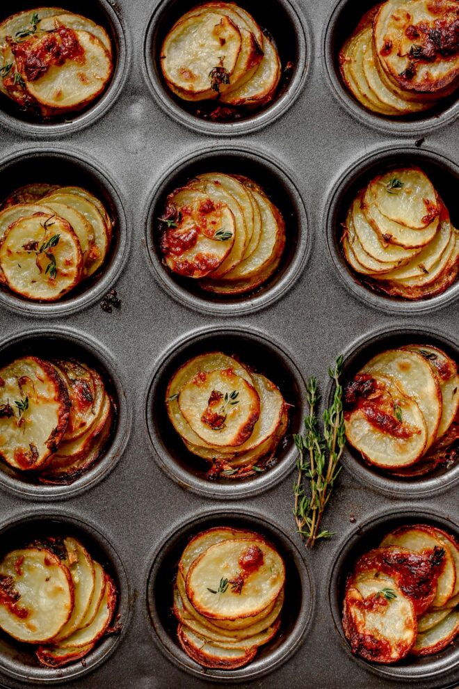 This is an overhead vertical image of a grey muffin tin. In the muffin tin cups are baked stacks of sliced potatoes. The potatoes have fresh thyme leaves on them. The muffin tin sits on a light grey surface.