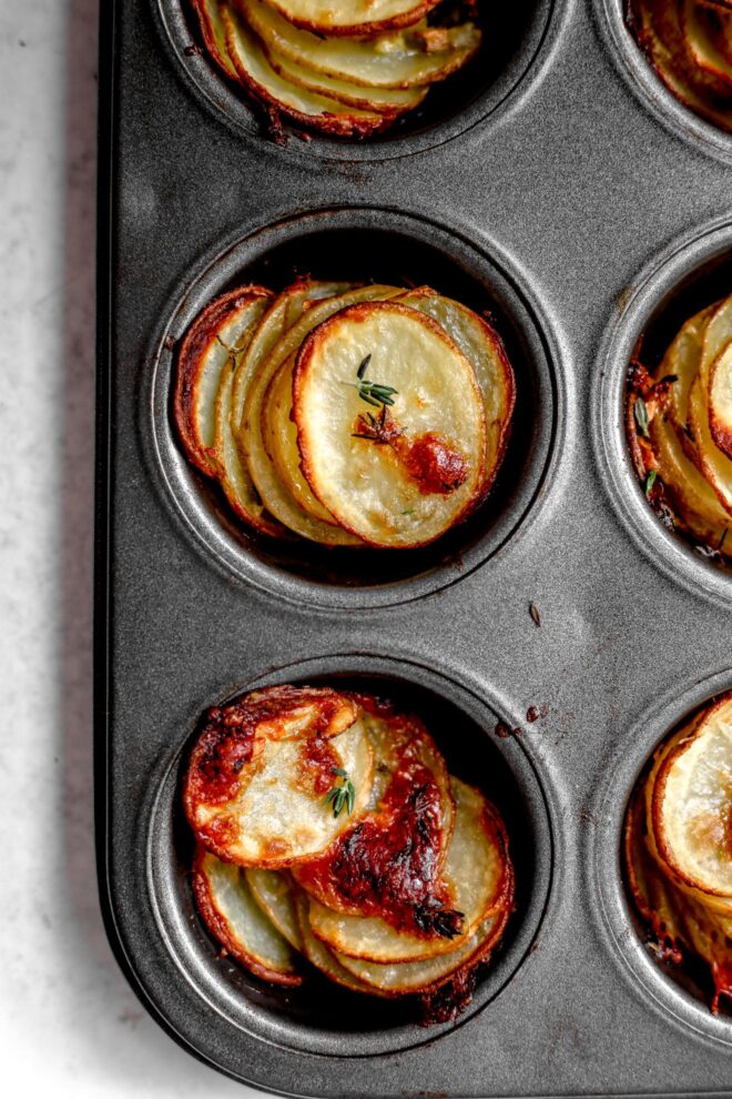 This is an overhead vertical image focusing on two cavities of a grey muffin tin. In the muffin tin cups are baked stacks of sliced potatoes. The potatoes have fresh thyme leaves on them. The muffin tin sits on a light grey surface.