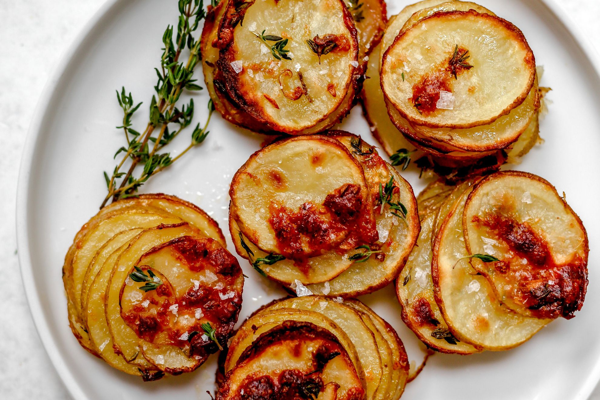 This is an overhead horizontal image of sliced stacked white potatoes. The potatoes have golden brown bubbles on them and are topped with flakey salt and fresh thyme leaves. The potato stacks are on a white plate on a white surface.