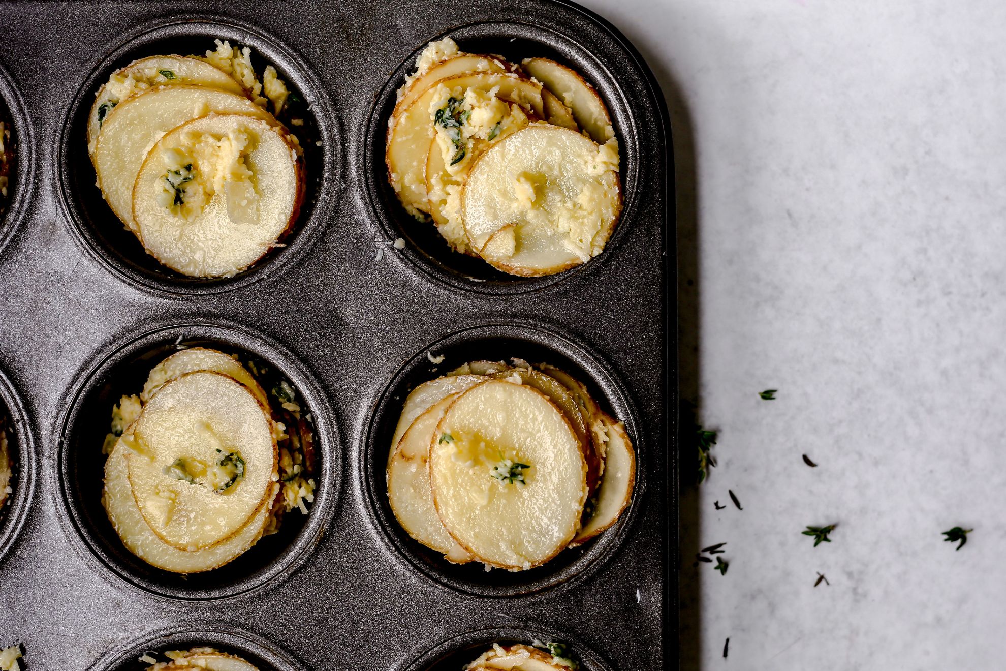 This is an overhead horizontal image of a grey muffin tin. The image focuses on the four corner muffin cups of the tin. In the muffin tin cups are stacks of sliced raw potatoes. The potatoes have fresh thyme leaves, butter, cheese and garlic on them. The muffin tin sits on a light grey surface.