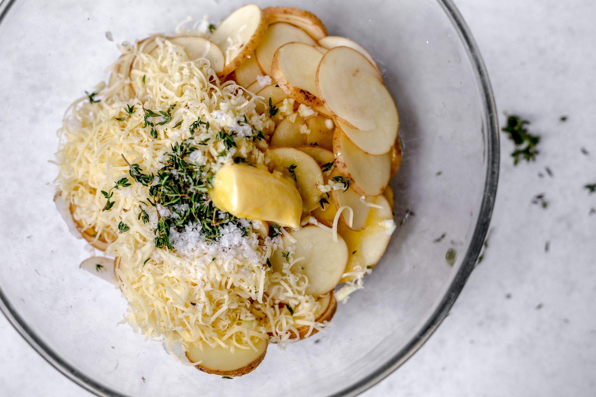 This is an overhead horizontal image of a glass bowl with sliced potatoes, shredded cheese, garlic butter, salt and fresh thyme leaves in it. The bowl sits on a light grey/white surface.
