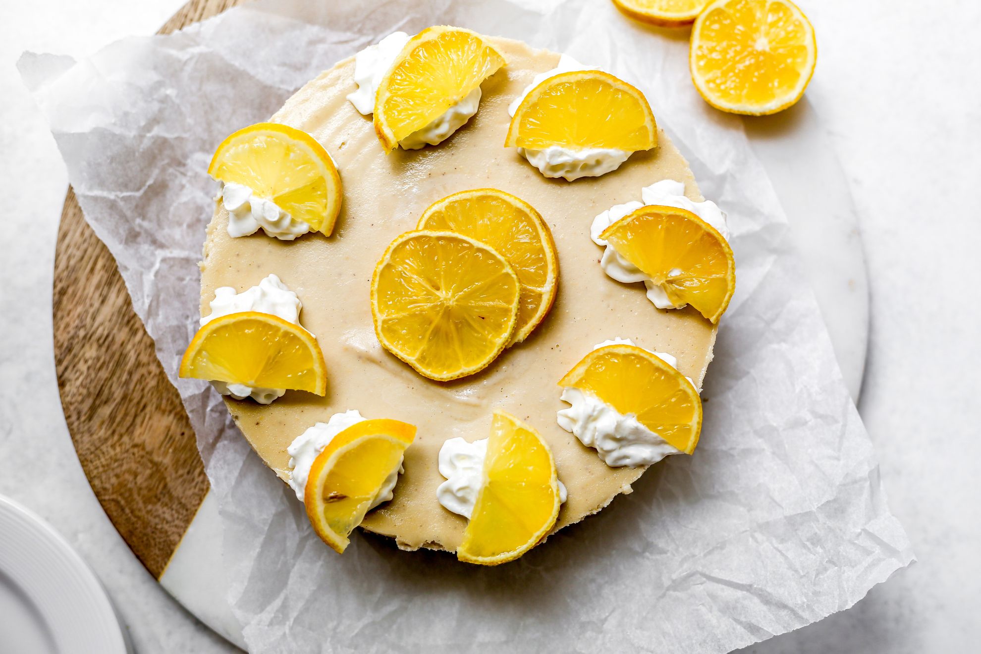 This is an overhead horizontal image of a no bake cheesecake on a piece of white parchment paper on a circular wood and marble cutting board. The cheesecake has dollops of whipped cream and lemon slices around the edge and two slices of lemon in the center. More lemon slices are next to the cheesecake to the top right, sitting on the cutting board. The cutting board sits on a white surface and a white plate is peaking into from from the bottom left corner.