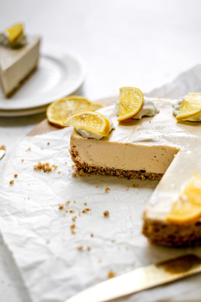 This is a vertical image looking at a no bake cheesecake from the side. The cheesecake is cut into and the image focuses on the nutty crust and creamy filling. The cheesecake is topped with dollops of whipped cream and wedges of lemon. The cheesecake sits on a white piece of parchment paper with some lemon wedges in the background and a stack of two white plates with a slice of cheesecake on top.