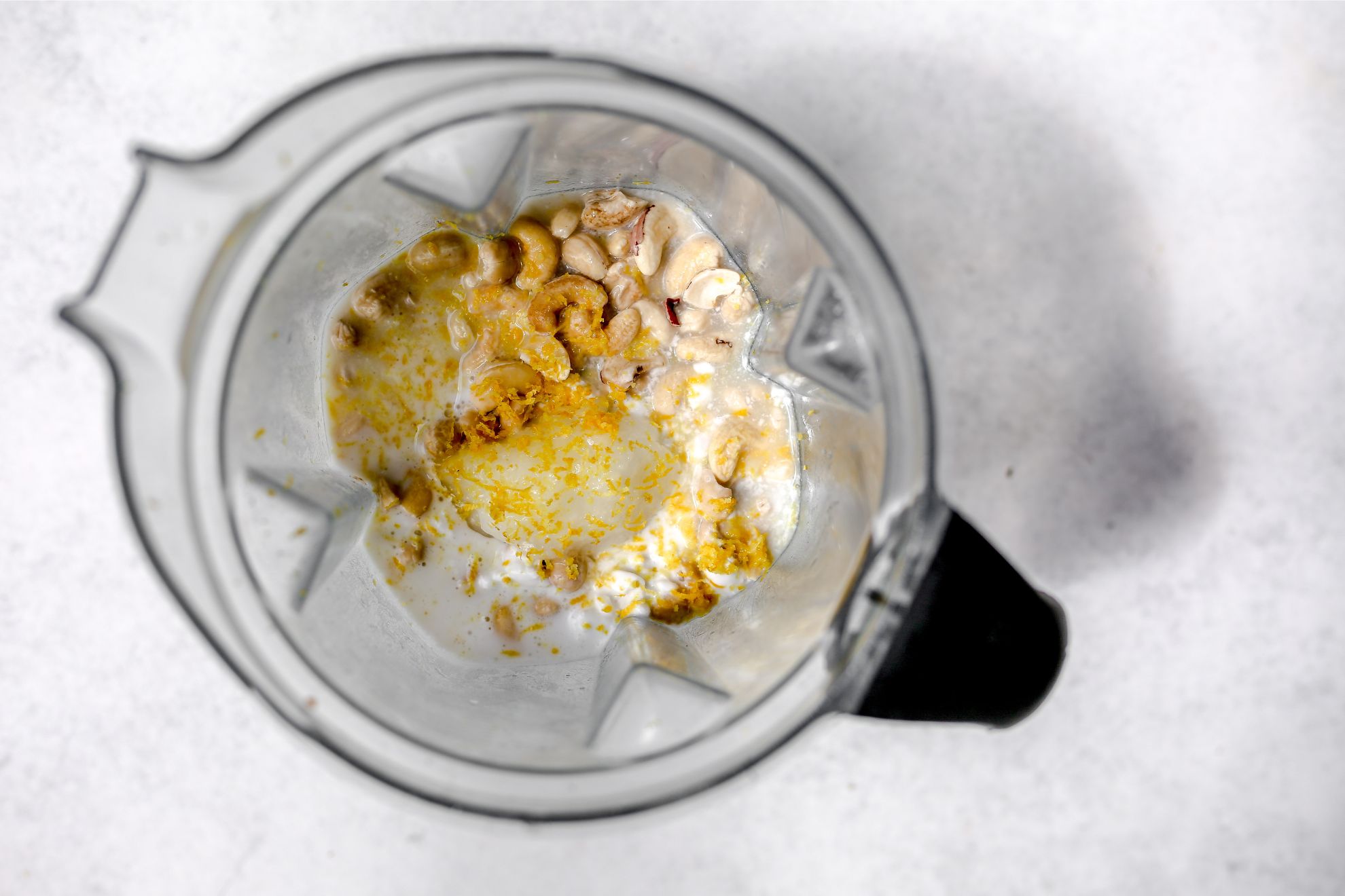 This is an overhead horizontal image looking into a blender. The blender sits on a white surface with ingredients in it. The ingredients appear to be cashews, milk, and lemon zest.