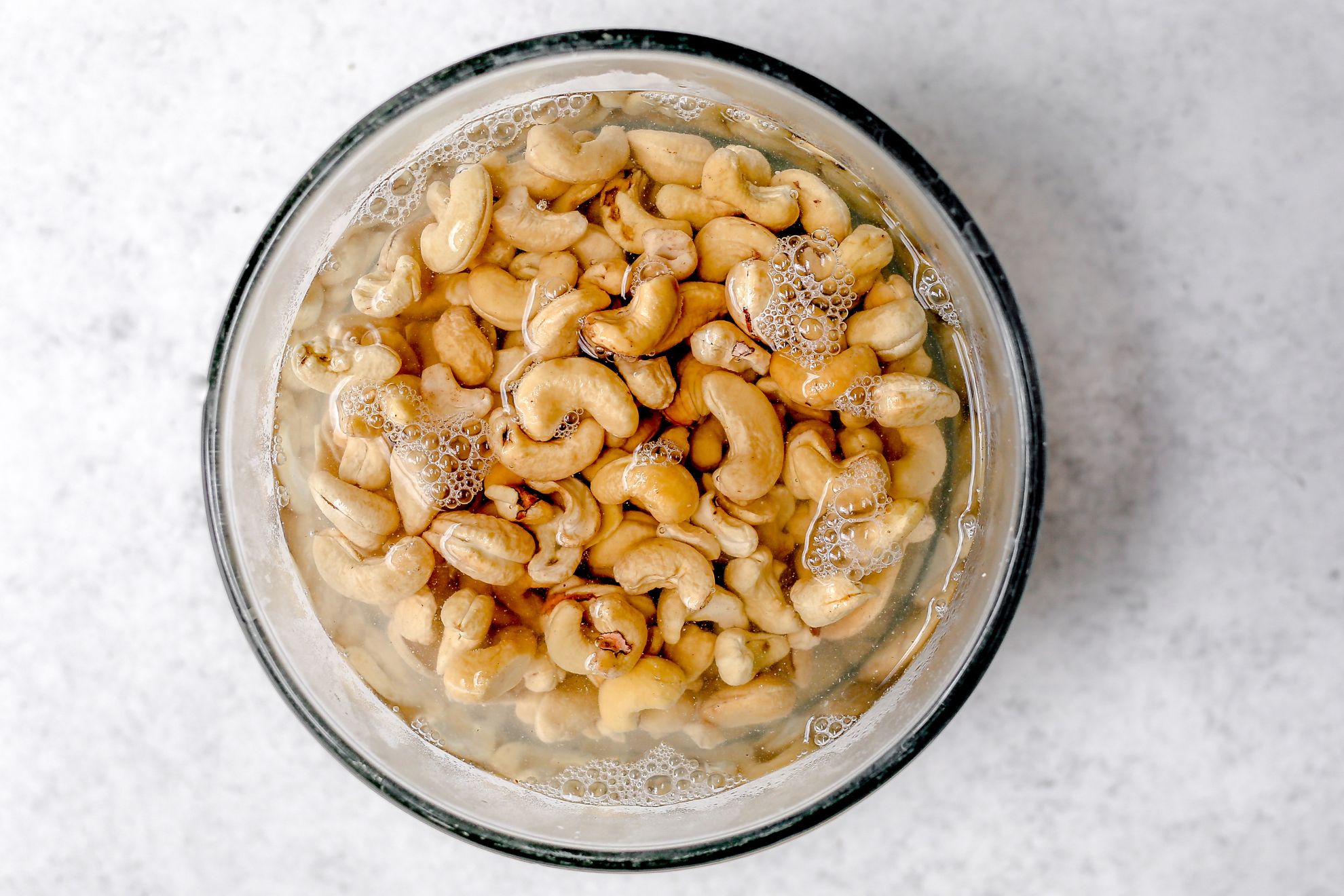 This is an overhead horizontal image of cashews soaking in water in a glass bowl. The bowl sits on a light grey surface.