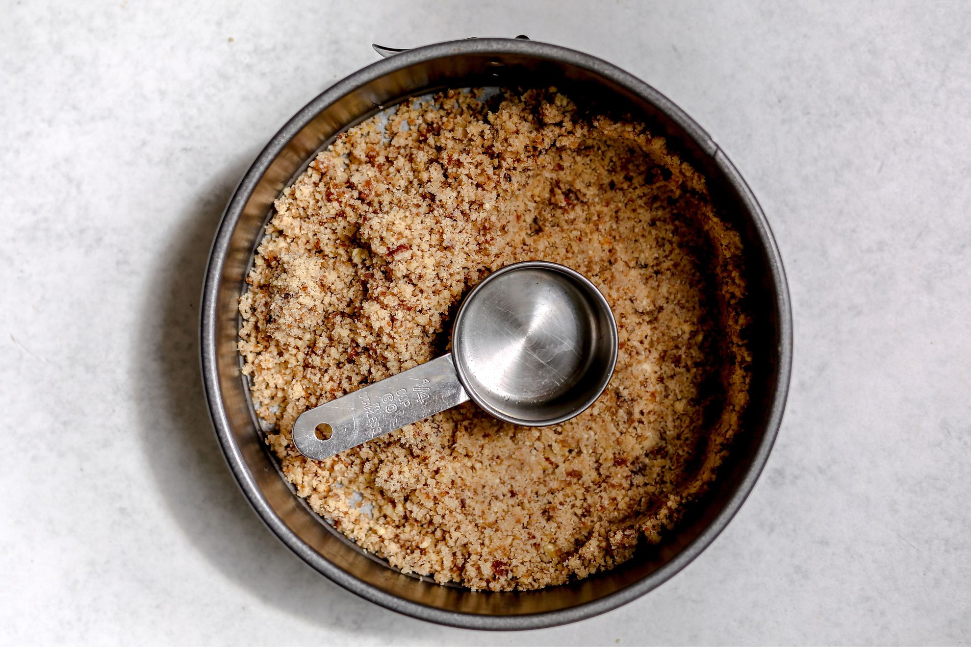 This is an overhead horizontal image of a circular pan with a nutty crumble crust at the bottom. A 1/4 cup dry measuring cup is in the center of the crust. It appears the right side of the crust has been pressed and flattened into the bottom and sides of the pan while the left side is still loose and crumbly. The pan sits on a white surface.