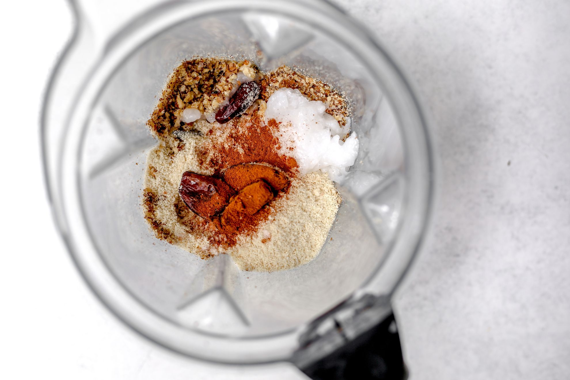 This is an overhead horizontal image looking into a blender. The blender sits on a white surface with ingredients in it. The ingredients appear to be walnuts, almond flour, dates, coconut oil, and cinnamon.