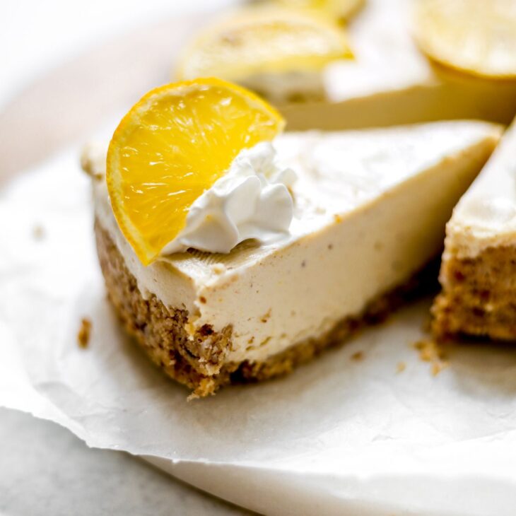 This is a vertical image looking at a no bake cheesecake from the side. The cheesecake is cut into and the image focuses on one slice pulled away from the rest of the cake. The slice has a nutty crust and creamy filling. The cheesecake is topped with dollops of whipped cream and wedges of lemon. The cheesecake sits on a white piece of parchment paper on a marble and wood circular cutting board on a white surface.