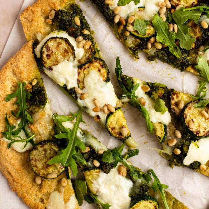 This is a vertical overhead image of a a few triangle slices of flatbread dolloped with pesto and shredded cheese, browned zucchini coins, burrata cheese, arugula and toasted pine nuts. The flatbread sits on a white piece of parchment paper on a light pink surface.