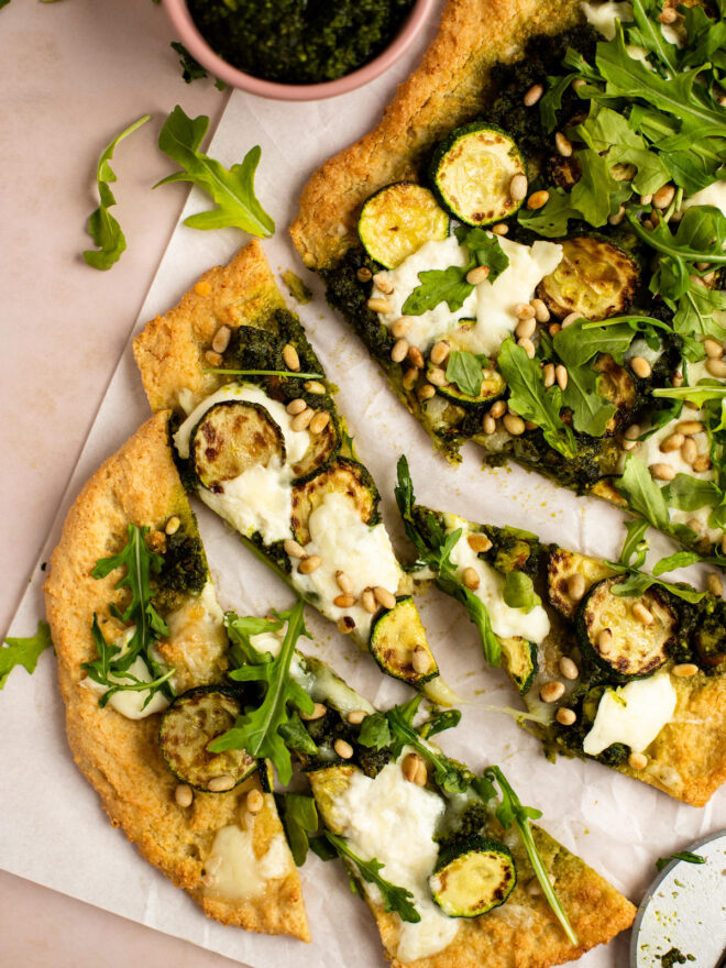 This is a vertical overhead image of an oval shaped crust dolloped with pesto and shredded cheese, browned zucchini coins, burrata cheese, arugula and toasted pine nuts. The flatbread sits on a white piece of parchment paper on a light pink surface and a few triangular slides are cut from the crust. A small pink bowl filled with pesto is to the top left of the image.
